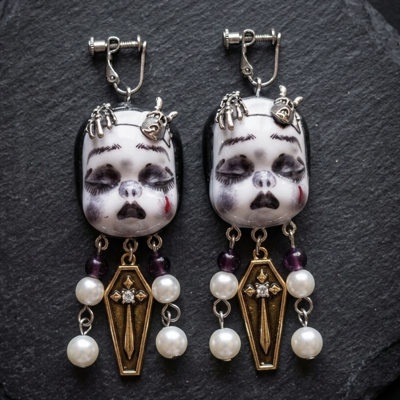 A pair of gothic dangle earrings featuring white porcelain doll faces with black painted details, silver-toned metal clips, and dangling coffin-shaped gold charms adorned with white pearls and dark purple beads, displayed on a dark slate background.