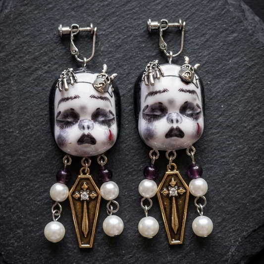 A pair of gothic dangle earrings featuring white porcelain doll faces with black painted details, silver-toned metal clips, and dangling coffin-shaped gold charms adorned with white pearls and dark purple beads, displayed on a dark slate background.